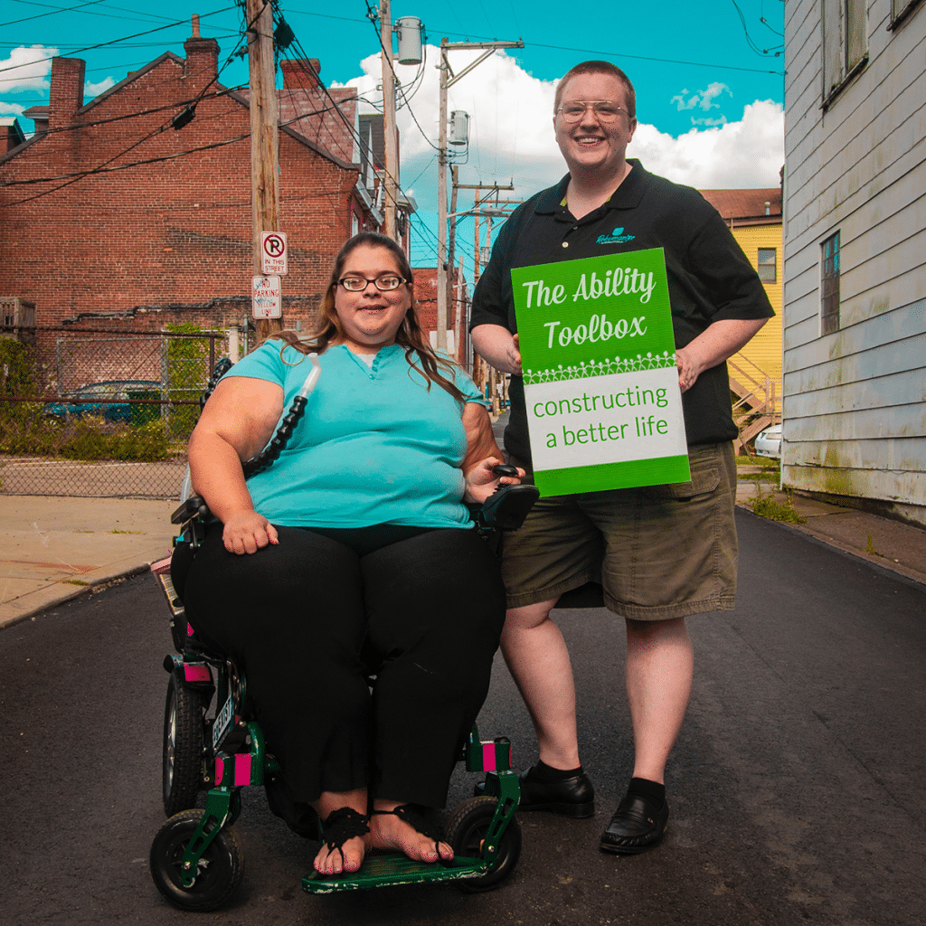 Woman in a wheelchair and friend holding sign that reads: The Ability Toolbox. Constructing a better life.