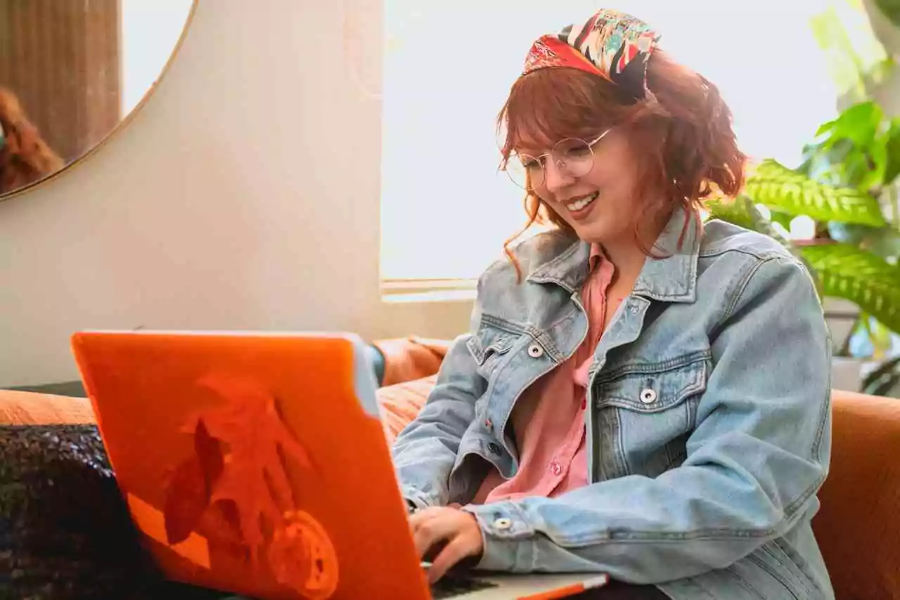 Woman with a disability working from home using a laptop on her sofa. She has red hair and is wearing a floral head scarf, denim jacket, and pink shirt.