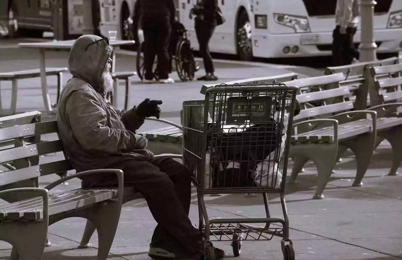 Homeless man with a beard sitting on a bench with a shopping cart holding his only worldly possessions.