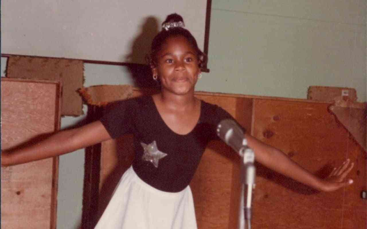 Alisa as a child, wearing a ballet dress and standing at a microphone.