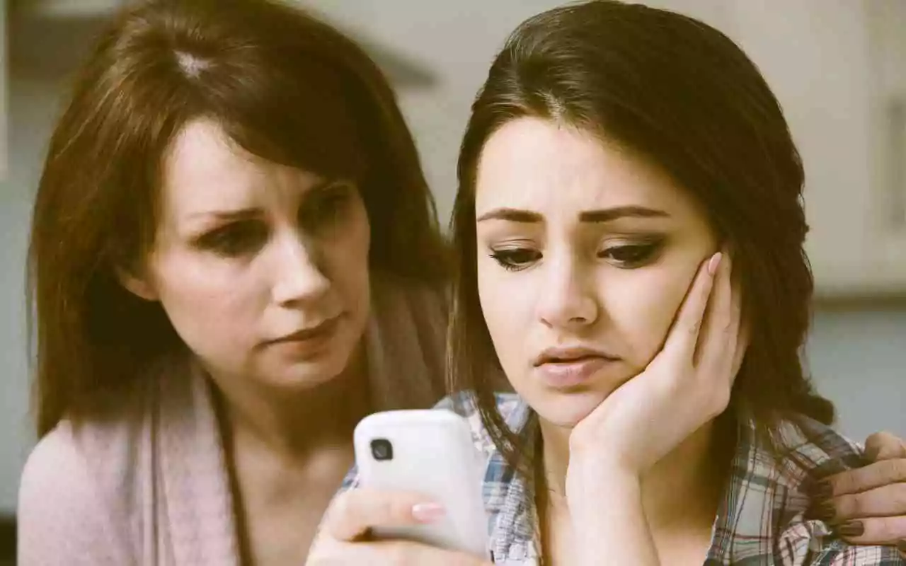 Teen feeling sad while looking at phone as mother looks on.