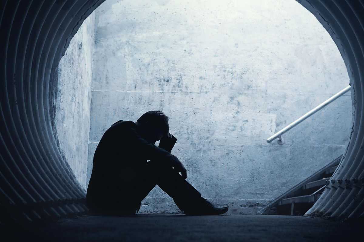 Man struggling with substance abuse and depression sitting at the base of a flight of stairs by a tunnel, holding his head in his hands.