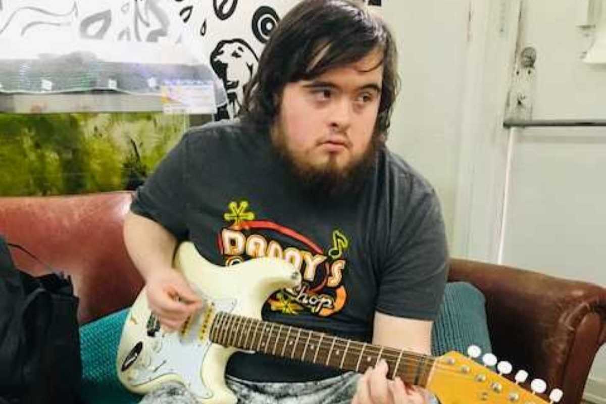Duncan Hines, a young man with a beard who has Down syndrome. He is sitting on a sofa and playing a white electric guitar.