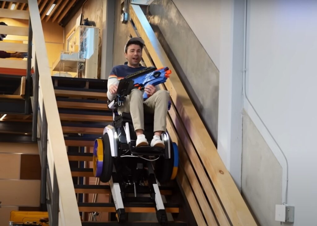 Mark Rober using a stair climbing wheelchair while holding a nerf gun.