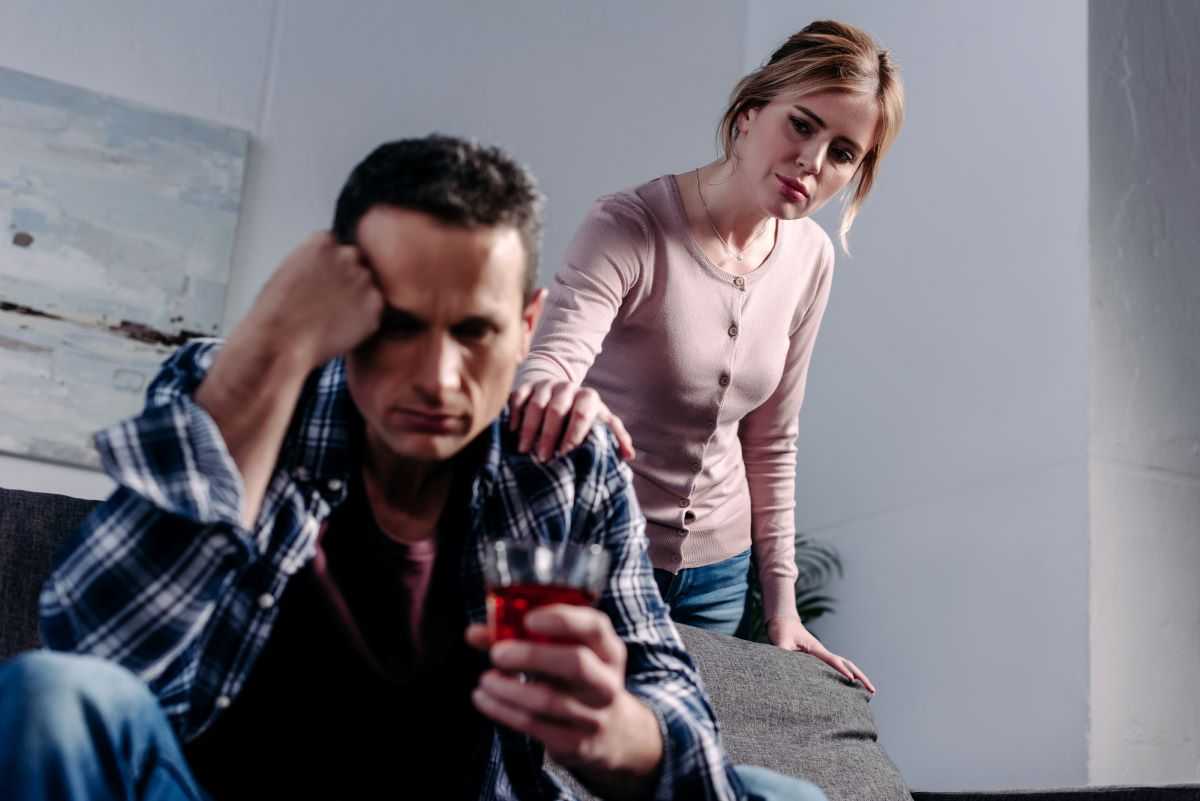 Man holding an alcoholic drink while his worried spouse places her hand on his shoulder.